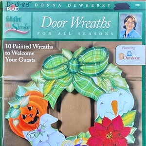May include: A book cover titled "Door Wreaths for All Seasons" by Donna Dewberry. The cover shows a painted wreath with a green plaid bow, a jack-o'-lantern, a snowman, a poinsettia, and Easter eggs. The book is from Plaid FolkArt.