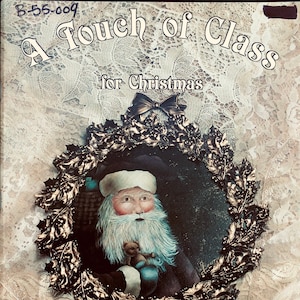 May include: A book cover titled "A Touch of Class for Christmas" featuring a painting of Santa Claus holding a teddy bear, framed by a gold-colored holly wreath. The background is a textured, off-white lace pattern.