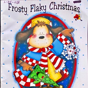 May include: A cartoon illustration of a dog wearing a red and white striped scarf and a blue jacket holding a small green Christmas tree. The dog is wearing brown boots and has a yellow hat. The illustration is titled "Frosty Flaky Christmas" and is by Annie Lang.
