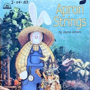 Puede incluir: Un libro titulado "Apron Strings" de Jayna James, que presenta un conejo de madera pintado con un mono y un sombrero de paja, sosteniendo una cesta de flores. El conejo está de pie en un entorno de jardín.