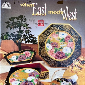 May include: A book cover with the title "When East Meets West" by Megumi Takada. The cover features a black and gold octagonal box with a floral design. The box is surrounded by other boxes and a glass jar with a lid.