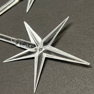 May include: Clear plastic star-shaped Christmas light bulb covers. The star has six points and a cylindrical base. Several are visible against a dark background. The star points are sharp and angular.