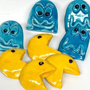 May include: A collection of ceramic Pac-Man and ghost figures. The ghosts are blue with black eyes and expressive faces. The Pac-Man figures are yellow with a black dot. The figures are arranged on a white surface.