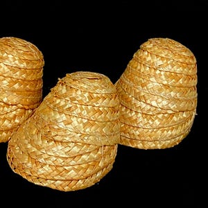Pu&ograve; includere: Tre cappelli di paglia intrecciata di forma conica sono esposti su sfondo nero. I cappelli sono di colore giallo dorato e sembrano essere fatti di paglia fittamente intrecciata. I cappelli sono impilati e hanno una parte superiore arrotondata.