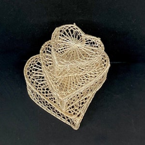 May include: Three woven heart-shaped baskets in varying sizes, stacked together. The baskets are a light beige color and have an open, airy design. The background is a solid black.
