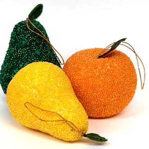 May include: Three fruit-shaped ornaments covered in tiny beads. One is a dark green pear, one is a bright yellow pear, and one is a round orange fruit. Each ornament features a small green fabric leaf and a thin gold hanging string attached to the top.