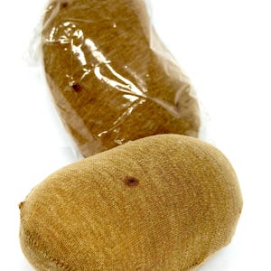May include: Two oval-shaped, tan-colored fabric objects with a soft, textured surface. One object is enclosed in a clear plastic bag, while the other is positioned in front, showing a small dark brown spot on its upper surface and visible stitching.