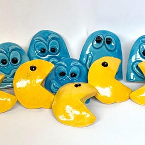 May include: A collection of ceramic Pac-Man and ghost figures. The ghosts are blue with black eyes, and the Pac-Man figures are yellow with black dots. These retro game-themed decorations are arranged on a white surface.