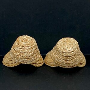May include: Two small, woven straw hats with a rounded crown and a wide brim. The hats are a light tan color and appear to be handmade. The hats are displayed against a dark background.