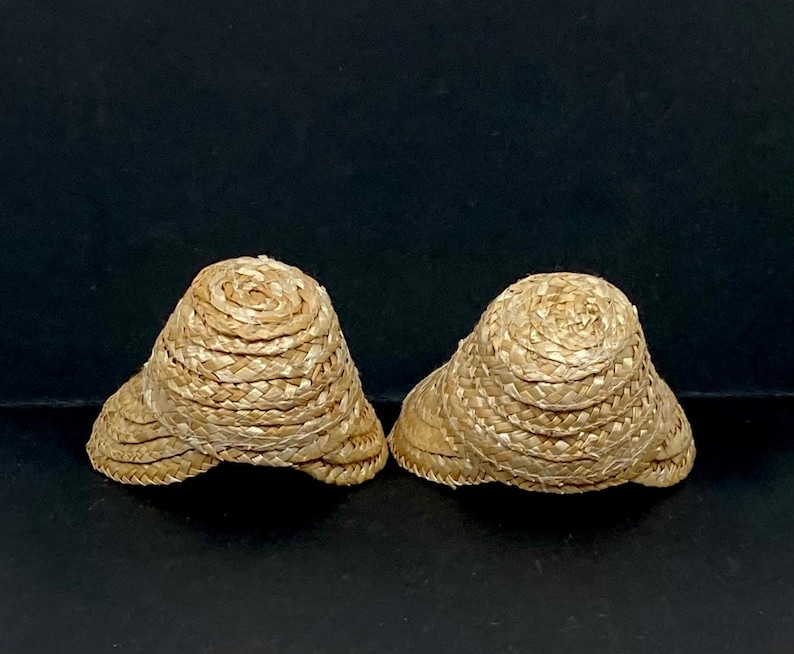 May include: Two small, woven straw hats with a rounded crown and a wide brim. The hats are a light tan color and appear to be handmade. The hats are displayed against a dark background.