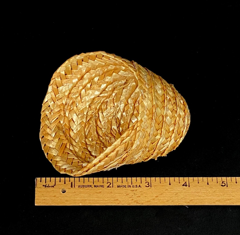 Pu&ograve; includere: Un piccolo cappello di paglia intrecciata di colore beige naturale. Il cappello &egrave; fatto di paglia fittamente intrecciata e ha una forma arrotondata. Un righello di legno &egrave; posto accanto al cappello.