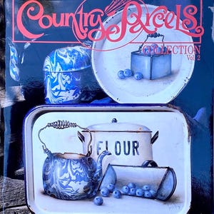 Op de afbeelding: Illustratie van keukenartikelen in landelijke stijl. De afbeelding toont een blauw-witte gemarmerde theepot, een bloembak en een dienblad met bosbessen. De tekst "The Country Reels Collection Vol 2" is zichtbaar.