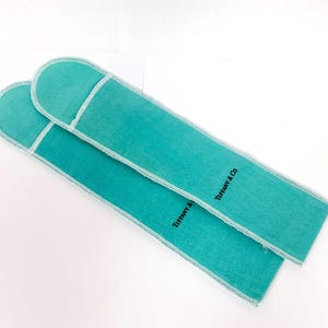 May include: Two rectangular, turquoise-colored pouches with rounded ends. The pouches are made of a soft material and have white stitching around the edges. The words "TIFFANY & Co." are printed in black on each pouch.