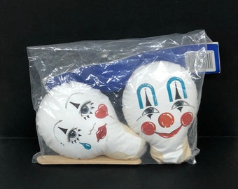 May include: Two white clown head fabric dolls with painted faces, packaged in a clear plastic bag. One clown has a red heart and tear, the other has a red nose and smile. The dolls are on a wooden tray.