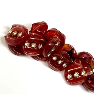 May include: A collection of vintage, dark red buttons with a glossy finish. Each button features a gold accent and three small, sparkling rhinestones. The buttons are strung together, suggesting they are part of a larger craft project or garment.