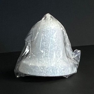 May include: A gray bell-shaped object, possibly a decorative item or a small sculpture, is encased in clear plastic. The bell has a textured surface and a wide base, tapering to a pointed top. The item is on a dark surface.