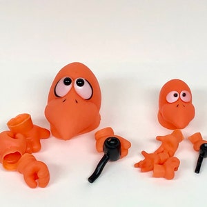 May include: Orange cartoon bird parts, including two heads, two hands, and two black-handled accessories, are arranged on a white surface. The bird heads have large, expressive eyes. The hands are in various poses.