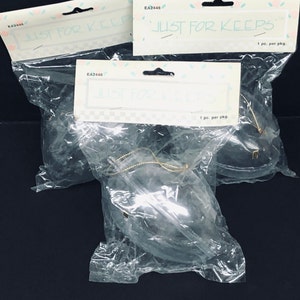 May include: Several clear plastic packages containing dark gray teardrop-shaped ornaments. Each ornament is packaged with a tag that reads "JUST FOR KEEPS". The ornaments have a gold string for hanging.