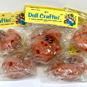 May include: Several 3" Pascal monkey faces with hands and feet, packaged in clear plastic. The faces are a reddish-brown color with black eyes and red accents. The packaging includes yellow labels with the text "Doll Craftin'" and "Made in Korea."
