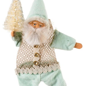 Puede incluir: Una caprichosa figura de gnomo navideño con un atuendo verde menta, sosteniendo un pequeño árbol color crema. El gnomo tiene una larga barba blanca, un gorro puntiagudo y un chaleco decorativo con campanas doradas. Los zapatos de la figura son negros.