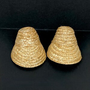 May include: Two small, woven straw hats with a flared brim and a rounded top. The hats are a light tan color and appear to be handmade. They are placed on a dark surface.