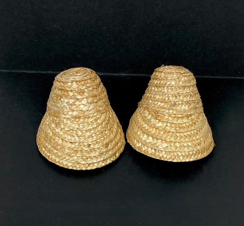 May include: Two small, woven straw hats with a flared brim and a rounded top. The hats are a light tan color and appear to be handmade. They are placed on a dark surface.