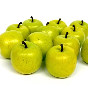 May include: A collection of twelve artificial apples in a bright lime green color. Each apple features a smooth, glossy finish with subtle dark speckles and a small, dark brown stem protruding from the top center of the rounded fruit shape.