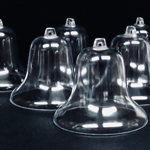 May include: Clear plastic bell-shaped ornaments with a small loop at the top. The ornaments have a flared base and a smooth, transparent surface. They are arranged on a dark surface, suggesting a holiday or craft theme.