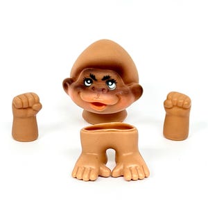 May include: A disassembled vintage troll doll with a brown head, arms, and legs. The troll has a smiling face with blue eyes and a brown hat. The arms and legs are separate from the body.