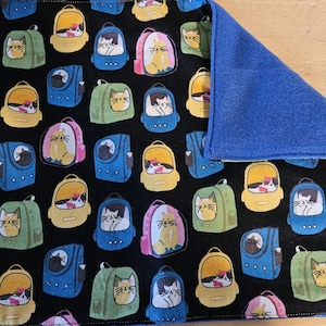 May include: A black fabric placemat with a repeating pattern of cats wearing backpacks. The backpacks are in various colors, including yellow, blue, green, and pink. The placemat has a blue fabric backing.
