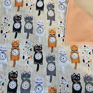 May include: A cream-colored fabric with a repeating pattern of black, orange, and white cats holding clocks. The fabric has a peach-colored fleece backing.