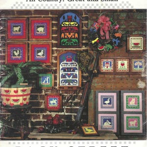 May include: A vintage book cover titled "SCHERENSCHNITTE" with the subtitle "All Country: Great and Small." The cover features framed cross-stitch designs of farm animals, houses, and trees. A decorative wreath and a basket with watermelon designs are also visible.