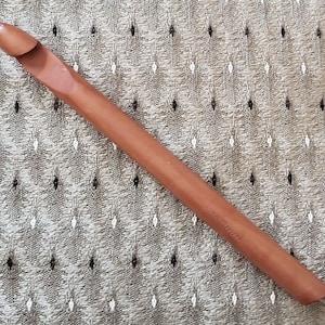 May include: A large, wooden crochet hook in a warm brown hue. The hook is angled at the tip and has a groove for catching yarn. The size is marked as 15 mm. The hook is lying on a patterned fabric.