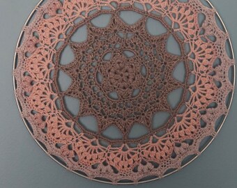 May include: A large, round, crocheted wall hanging with a pink and brown ombre design. The hanging is made with a delicate, lacy stitch and has a starburst pattern in the center.
