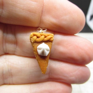 K&ouml;nnte beinhalten: Ein Miniatur-K&uuml;rbiskuchen-Anh&auml;nger mit einem wei&szlig;en Schlagsahne-Topping und einer geflochtenen Kruste. Der Anh&auml;nger ist aus Polymer Clay gefertigt und an einer silbernen Kette befestigt.