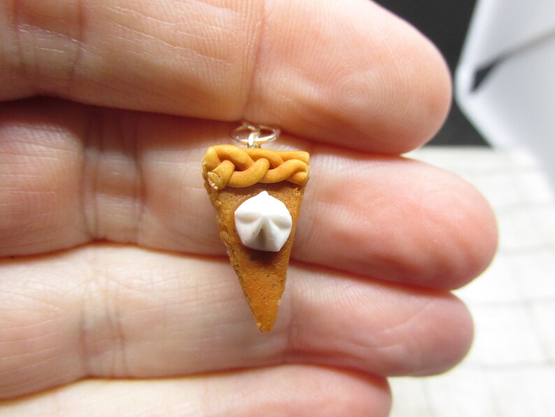K&ouml;nnte beinhalten: Ein Miniatur-K&uuml;rbiskuchen-Anh&auml;nger mit einem wei&szlig;en Schlagsahne-Topping und einer geflochtenen Kruste. Der Anh&auml;nger ist aus Polymer Clay gefertigt und an einer silbernen Kette befestigt.