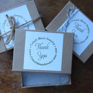 K&ouml;nnte beinhalten: Drei kleine braune Pappkartons mit wei&szlig;en Etiketten. Jedes Etikett hat einen Kreis mit dem Text "Cobalt Moon Jewelry" und "Thank You" darin. Die Kartons sind mit Schnur gebunden.