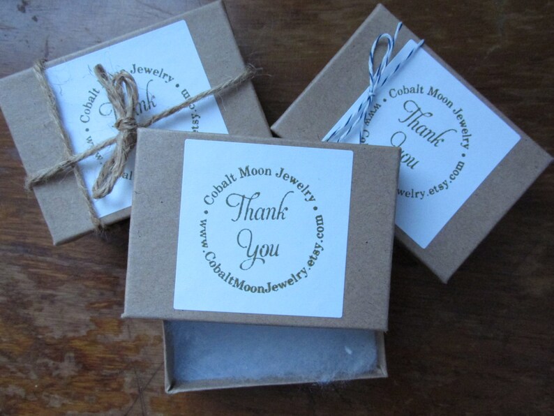 K&ouml;nnte beinhalten: Drei kleine braune Pappkartons mit wei&szlig;en Etiketten. Jedes Etikett hat einen Kreis mit dem Text "Cobalt Moon Jewelry" und "Thank You" darin. Die Kartons sind mit Schnur gebunden.