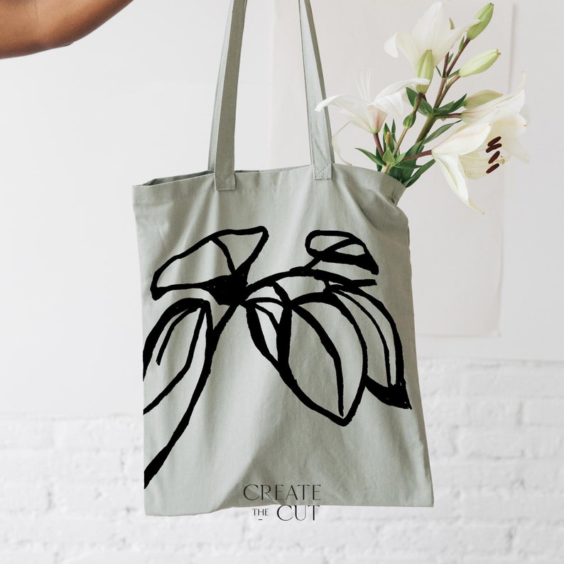 Op de afbeelding: Een lichtgroene tote bag met een zwarte lijntekening van een plant op de voorkant. De tas heeft de tekst "CREATE THE CUT" onderaan gedrukt.