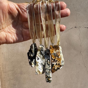May include: Five natural stone pendants with gold ribbon hanging loops. The stones vary in shape, color, and pattern, including black, white, brown, and gold. The pendants are held up by a hand.