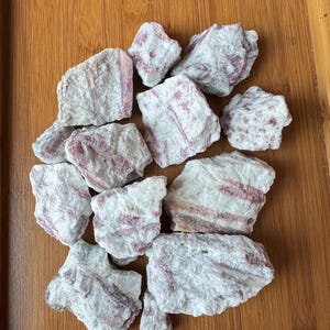 May include: A collection of rough-cut tourmaline stones, showcasing a mix of white and pink hues. The stones are irregularly shaped, with visible crystalline structures and pink streaks running through the white base. They are arranged on a wooden surface.