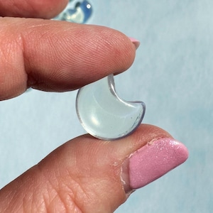 May include: A small, translucent, crescent moon-shaped glass bead. The bead is held between two fingers against a light blue background. The bead has a slight blue-green tint and is smooth.