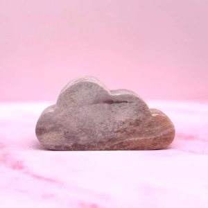 May include: A small, cloud-shaped stone sculpture in shades of grey and brown. The stone has a smooth, polished appearance and sits on a pink surface. The sculpture is a decorative item.