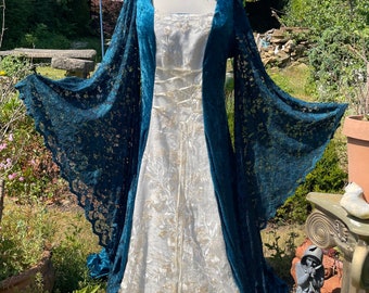 Puede incluir: Un vestido blanco con bordados dorados y una sobrecapa de terciopelo azul verdoso con mangas de encaje. El vestido está en un maniquí en un entorno de jardín.