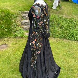 May include: A black velvet gown with a sheer overlay featuring embroidered floral details. The gown has a fitted bodice and a flowing skirt.