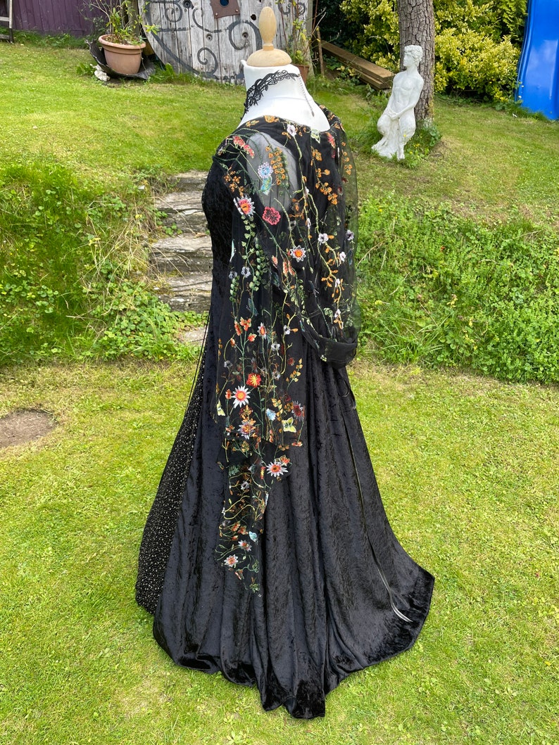 May include: A black velvet gown with a sheer overlay featuring embroidered floral details. The gown has a fitted bodice and a flowing skirt.