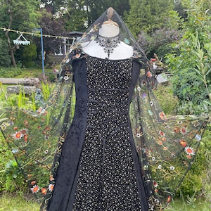 May include: A black velvet gown with a fitted bodice and a flowing skirt. The gown is adorned with small gold sequins and has a black lace overlay with floral embroidery. The gown is displayed on a mannequin with a black lace choker.