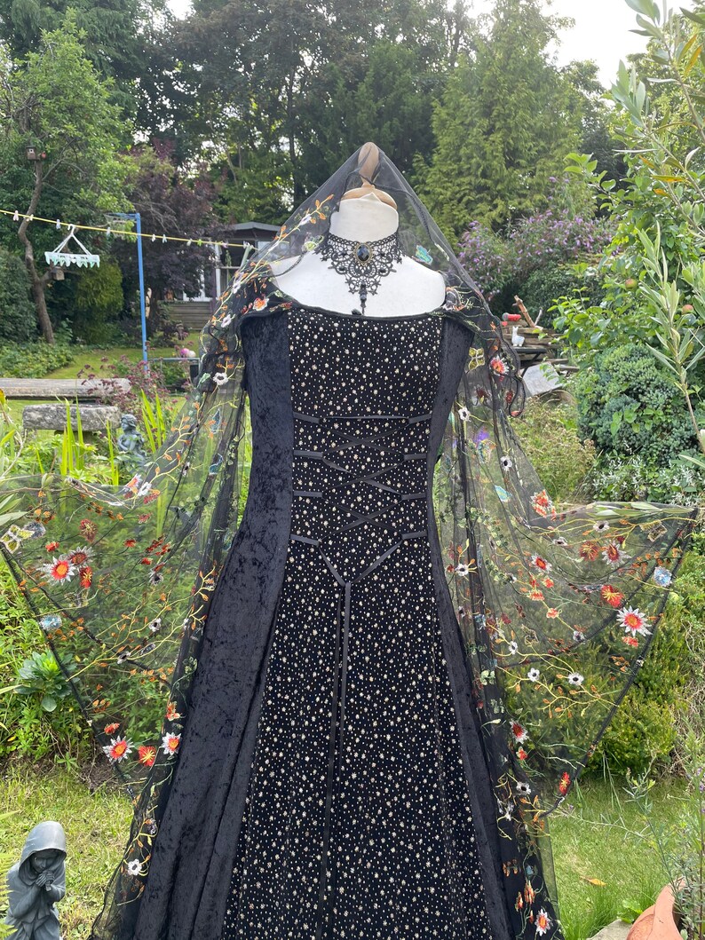 May include: A black velvet gown with a fitted bodice and a flowing skirt. The gown is adorned with small gold sequins and has a black lace overlay with floral embroidery. The gown is displayed on a mannequin with a black lace choker.