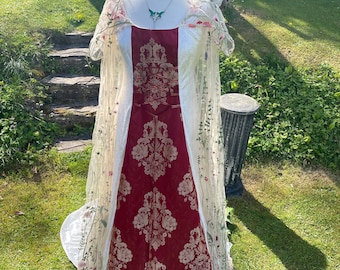 Puede incluir: Un vestido largo, rojo y blanco con una superposición de encaje y bordados florales. El vestido tiene un corpiño ajustado y una falda fluida. El vestido se muestra en un maniquí en un entorno de jardín.