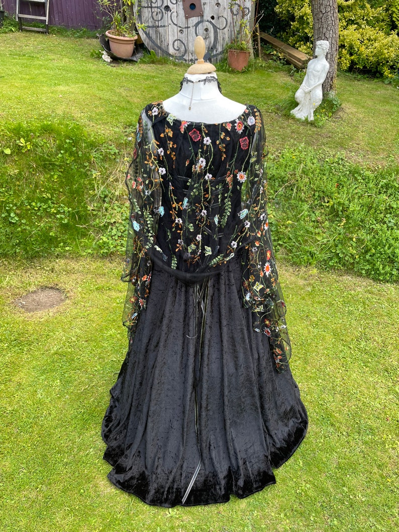 May include: A black velvet gown with a sheer overlay featuring floral embroidery in shades of orange, green, and white. The gown has a fitted bodice and a flowing skirt.